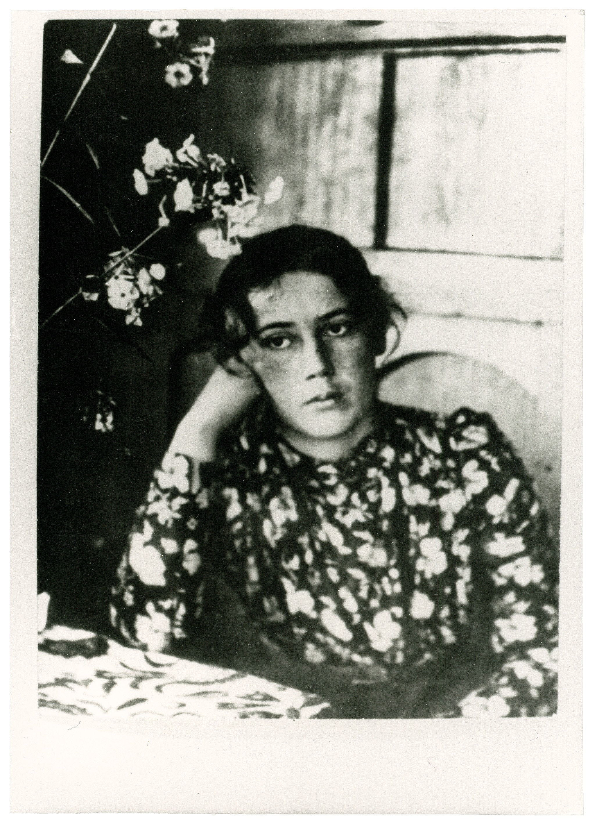 This black-and-white photograph shows Lisbeth Stern, née Schmidt (1870–1963), the younger sister of Käthe Kollwitz, as a young woman with dark, slightly tousled hair, seated thoughtfully at a table outdoors, presumably on a terrace. She wears a dark dress with a light, small-scale floral pattern and rests her head on her right hand, her expression conveying a sense of melancholy. Light-coloured blossoms appear in the left foreground, while the background is defined by a simple window and a textured wall.