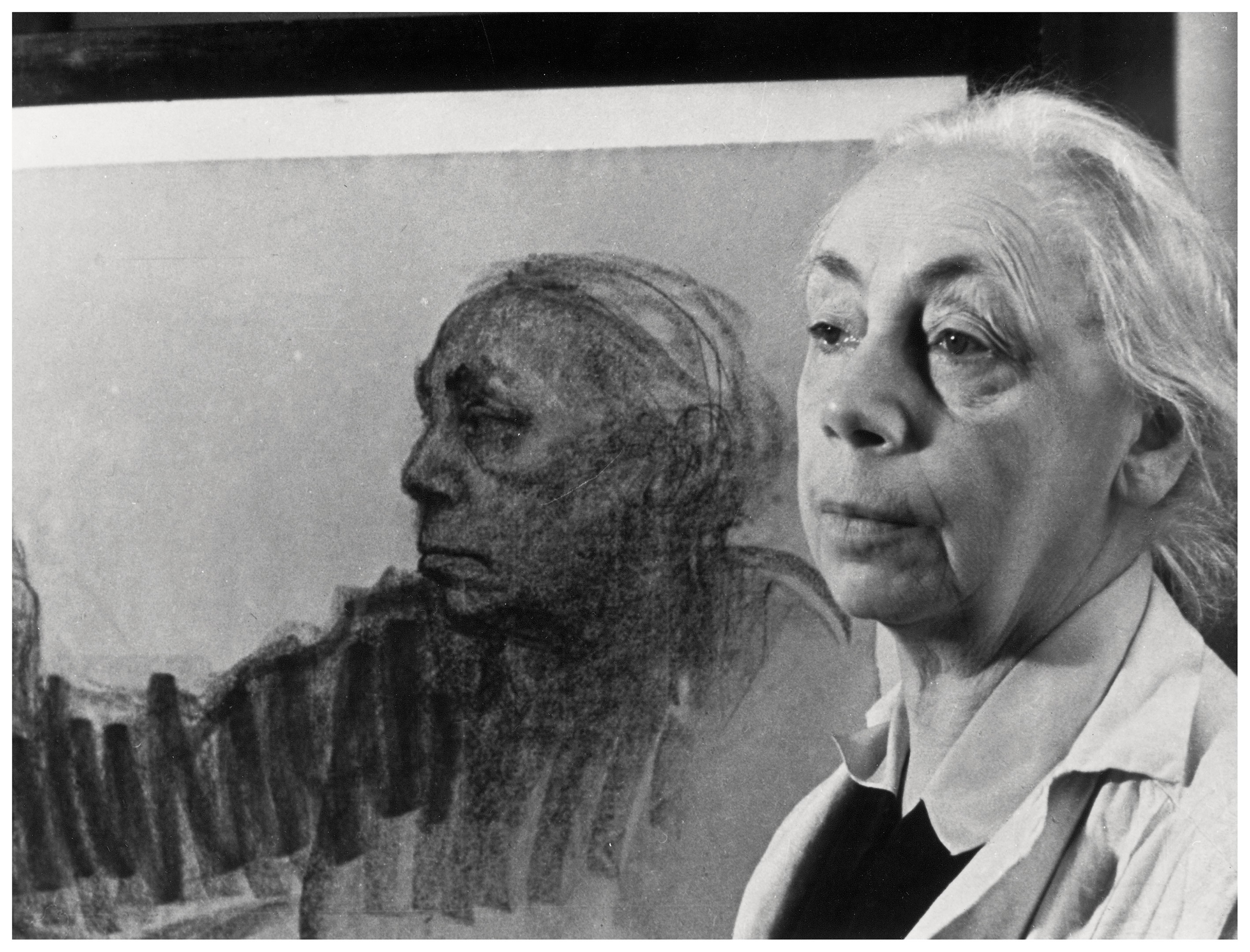 The landscape-format black-and-white photograph shows Käthe Kollwitz in the right foreground, while a life-size drawing of her own face in profile can be seen behind her on the left. She is wearing a light-colored work jacket over dark clothing and gazes slightly past the viewer with a serious, contemplative expression.