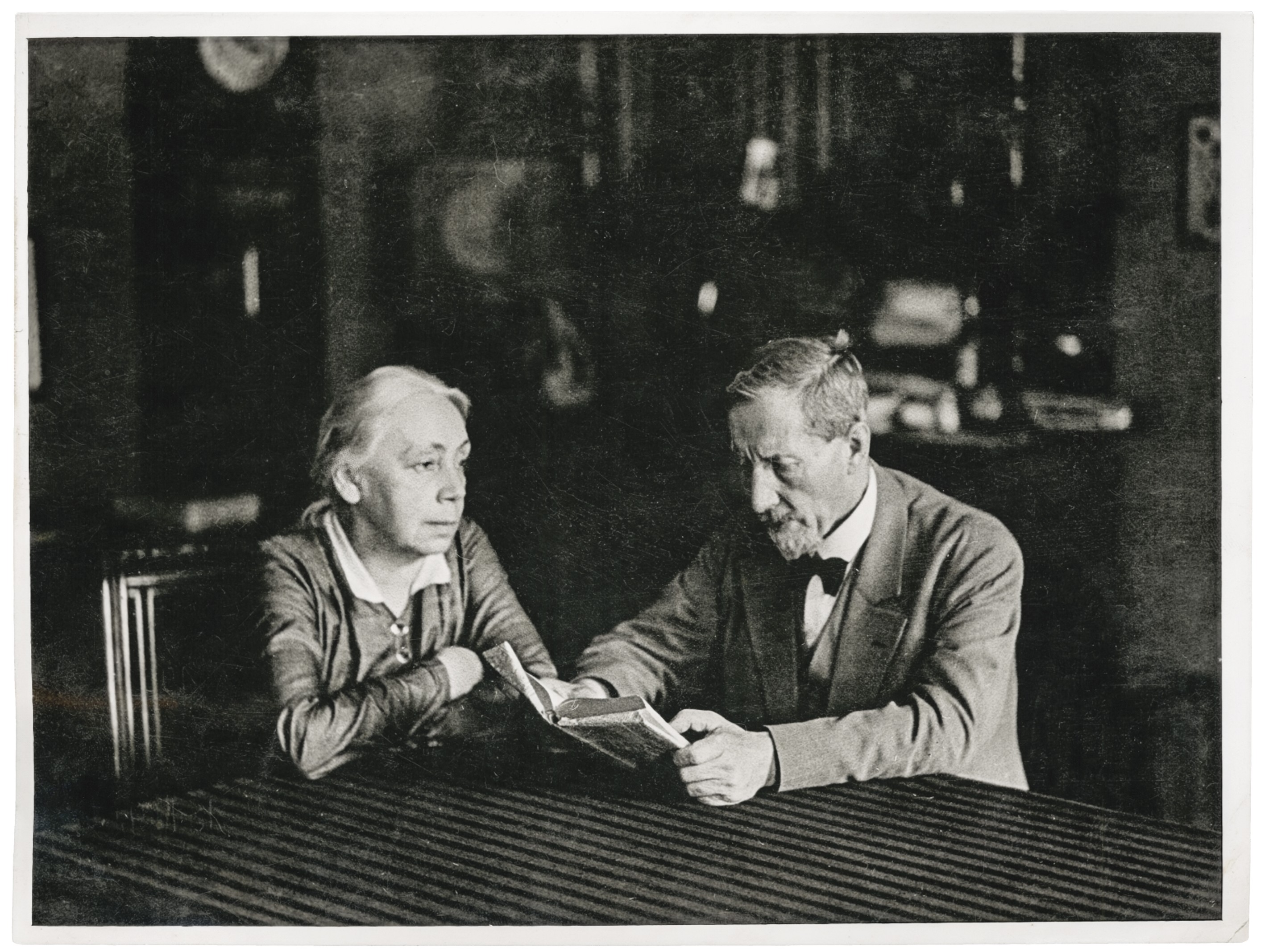 The black-and-white photograph by Erich Salomon from 1931 shows the artist Käthe Kollwitz and her husband Karl seated at a table in a middle-class living room. Karl, wearing a suit and a beard, holds a small book in his hands and appears to be reading aloud from it. Käthe sits opposite him with her arms folded on the tabletop, looking at her husband attentively and calmly.