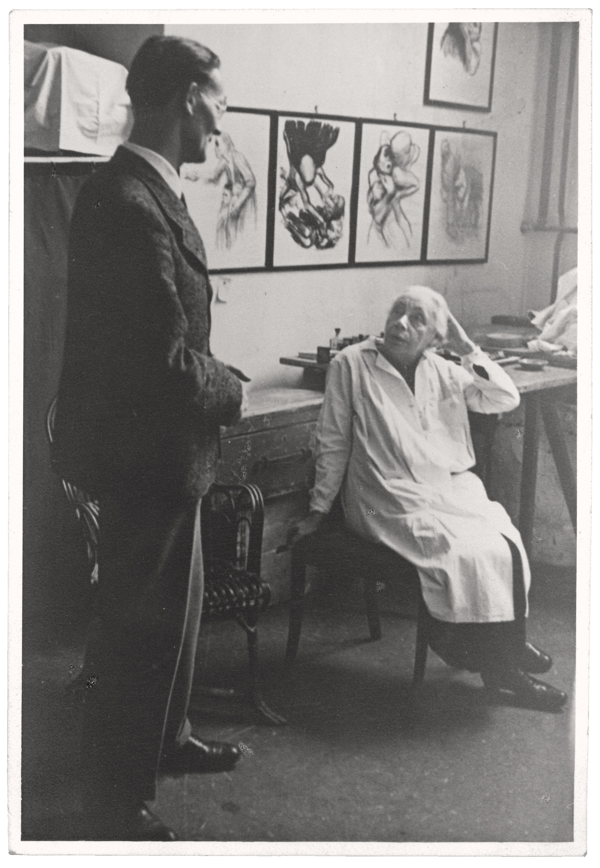 Black-and-white photograph of Käthe Kollwitz in her studio. She is seated on a chair wearing a light-colored smock and looking up at a standing man in a dark suit. Several framed drawings are visible on the wall behind them, and a worktable with papers and materials can be seen in the background. The studio appears modest and softly lit.