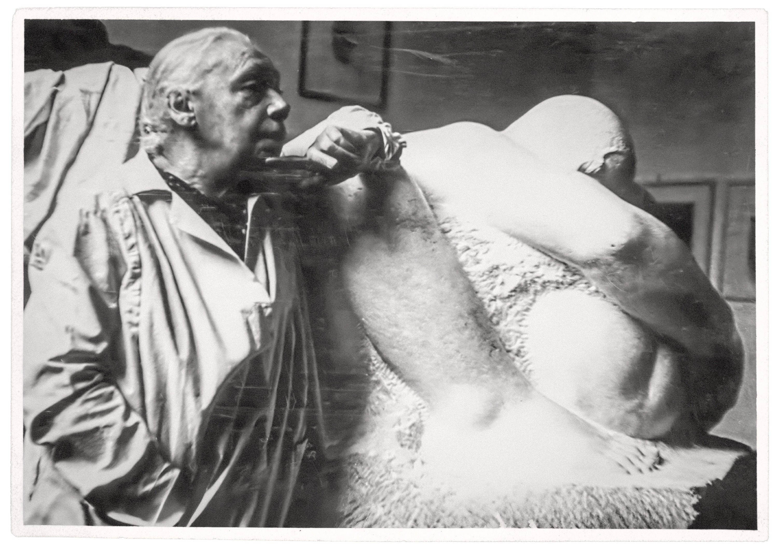 Black-and-white photograph of Käthe Kollwitz leaning her arm on a large sculpture of a mother embracing two children in her studio. She wears a light-colored work coat and looks to the side with a thoughtful expression. The sculpture fills much of the image, with framed artworks and studio elements faintly visible in the background.