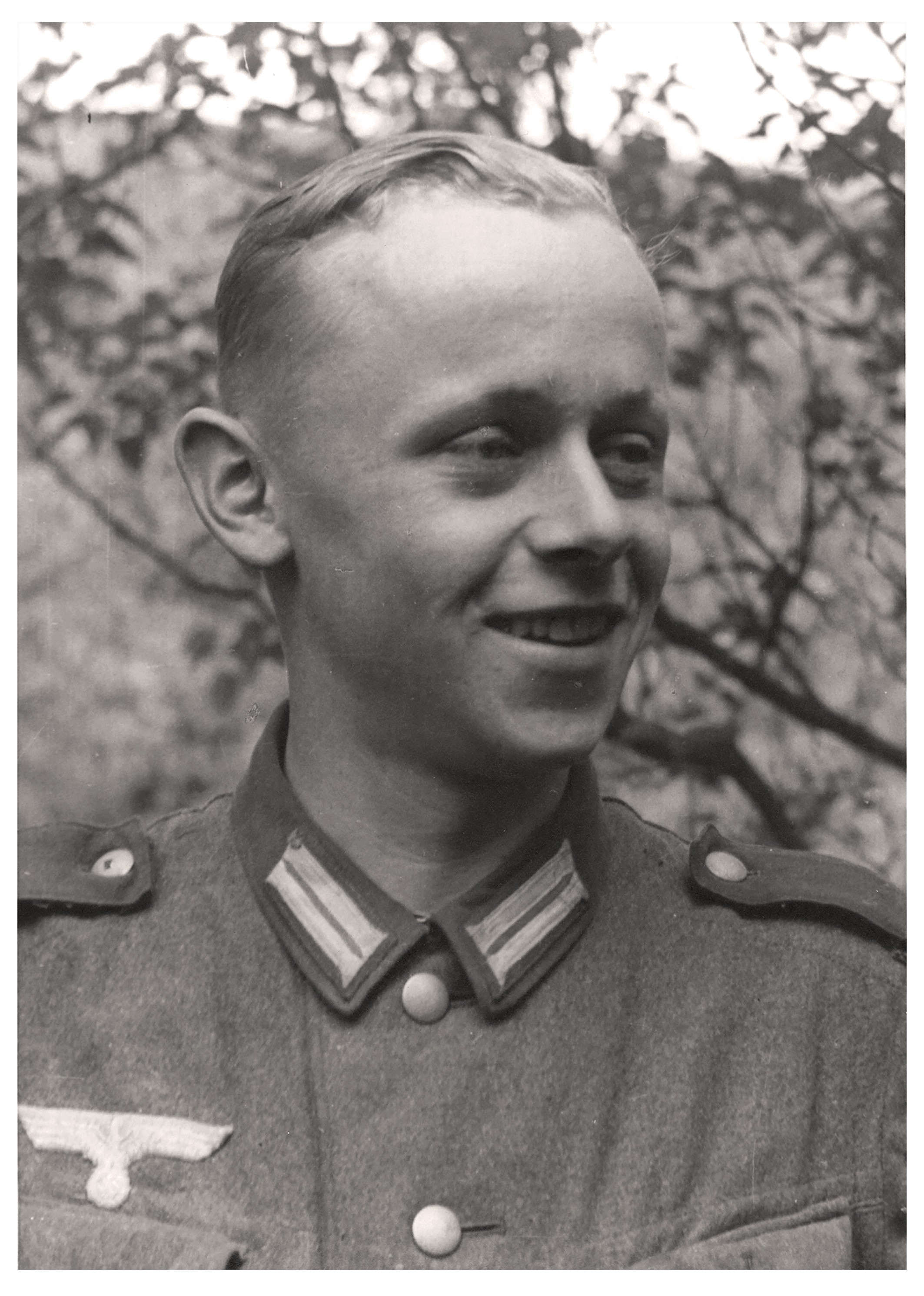 The black-and-white portrait from 1941 shows a young, smiling man wearing a German Wehrmacht uniform from the Second World War. He is dressed in a field tunic with distinctive collar insignia and the national emblem positioned above the right breast pocket, while his gaze is directed to the side. His light-colored hair is neatly combed to one side, and the background consists of slightly blurred leaves and branches outdoors.