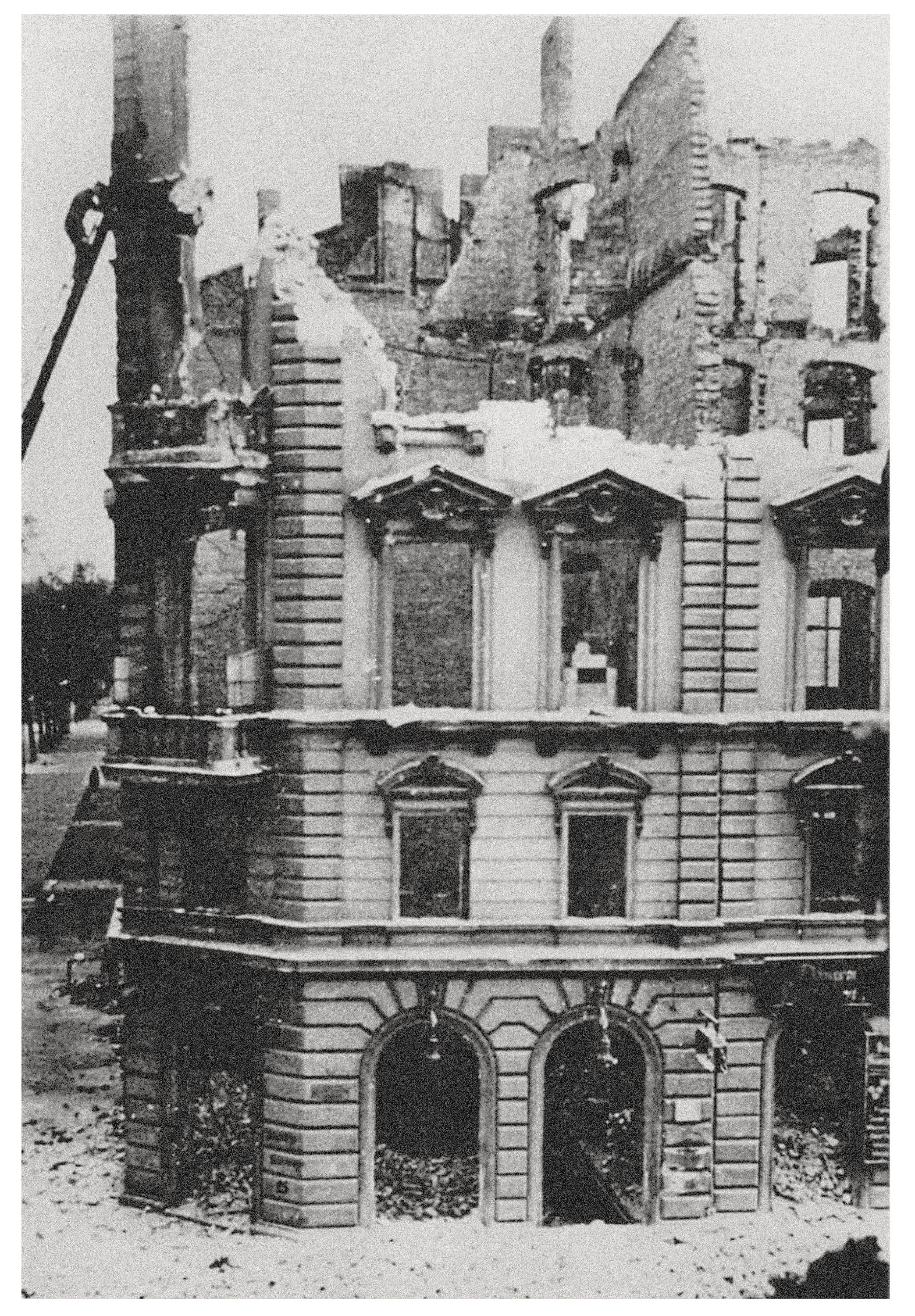 This black-and-white photograph shows the bombed-out ruin of a magnificent, multi-storey building with a neoclassical façade, of which only the outer walls still stand. The window cavities are empty, the roof is missing entirely, and inside the floors have collapsed, so that one looks through the façade into the open sky. On the ground lie heaps of rubble before the surviving round-arched entrances at ground-floor level.