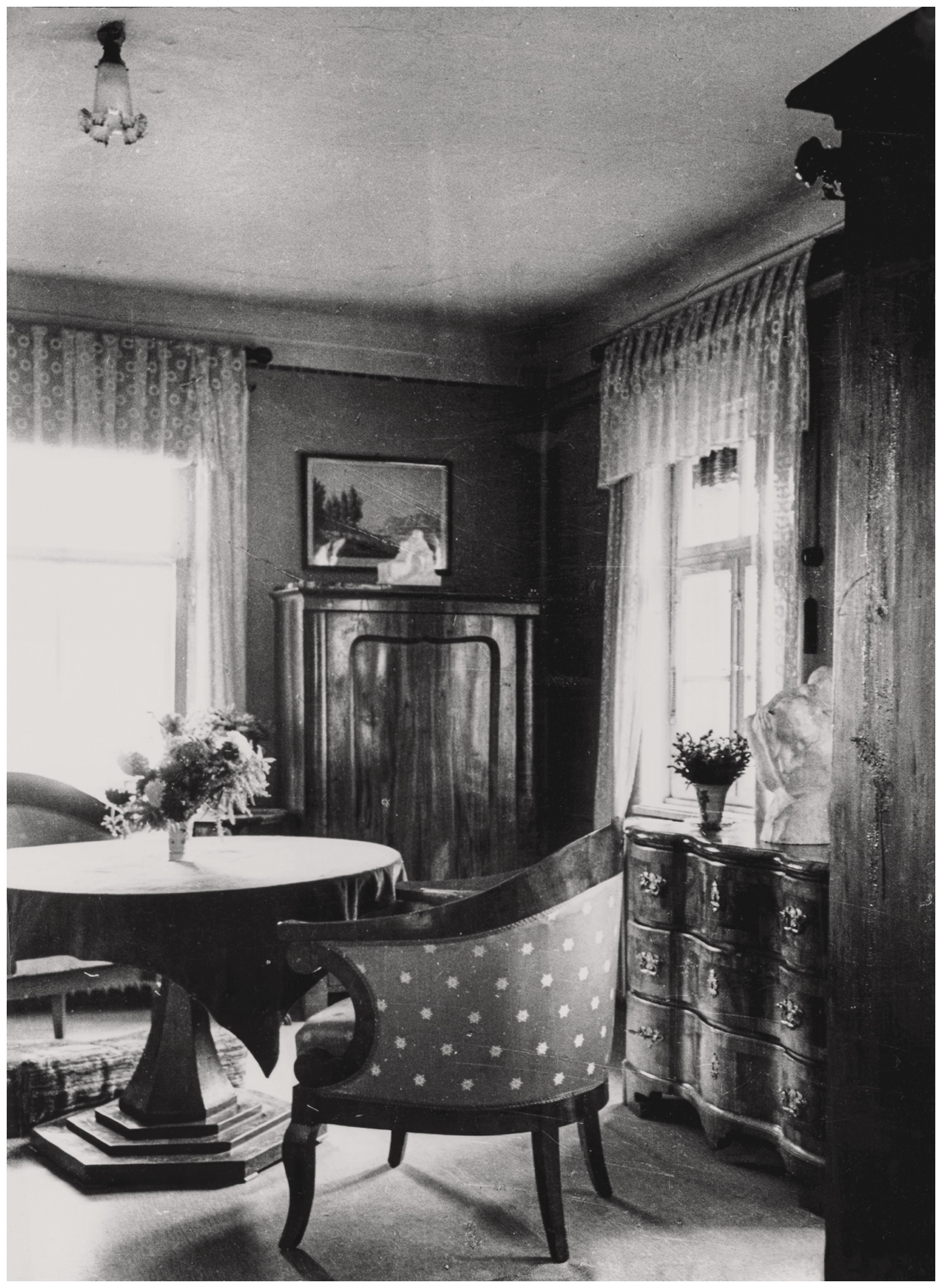 This black-and-white photograph shows a cozily furnished room with a round table in the center, surrounded by upholstered armchairs with a dotted pattern. In the background stand dark, classical wooden furnishings, including a curved chest of drawers and a cupboard, while light lace curtains at the windows fill the room with soft light. Vases with flowers and small sculptures are arranged on the furniture, lending the room a personal yet museum-like atmosphere: these are the plaster models created by Käthe Kollwitz—Two Waiting Soldiers’ Wives on the cupboard and Mother with Child over Her Shoulder on the chest of drawers to the right.