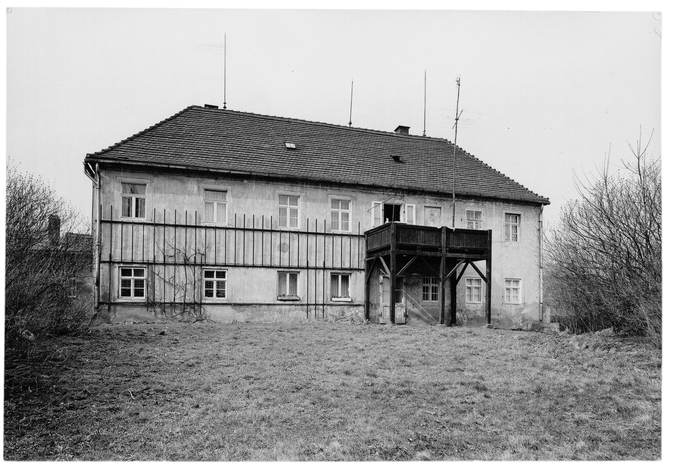 The black-and-white photograph shows the rear view of a large, two-story residence with a high hipped roof, situated on an expansive grassy property. Particularly striking is a massive dark wooden balcony at the center of the façade, supported by tall posts above the entrance area. The light-colored plaster façade is строго structured by a horizontal timber framework and numerous evenly arranged mullioned windows.