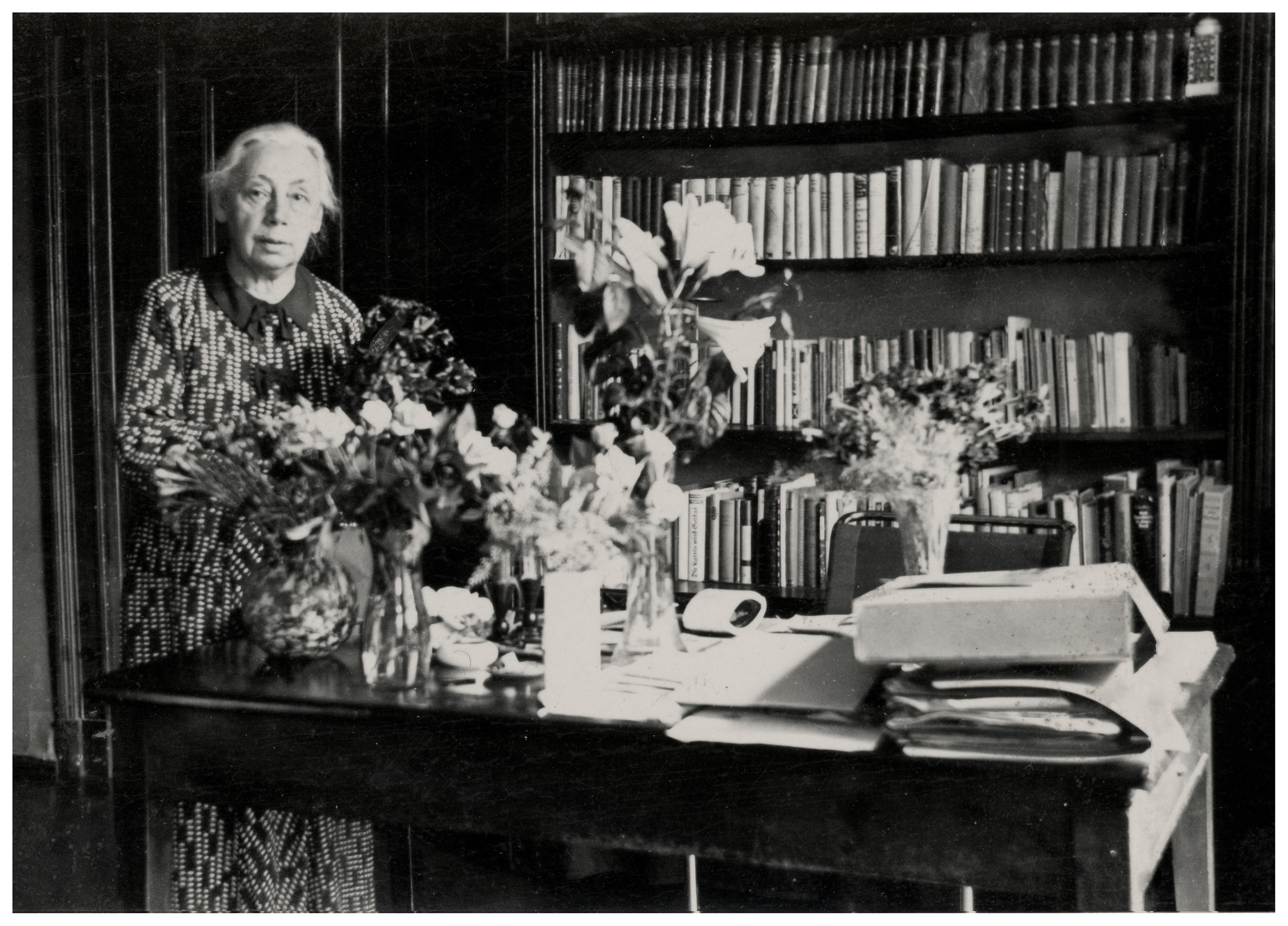 Black-and-white photograph of Käthe Kollwitz standing in a room with light-colored hair tied back. She is positioned in front of a large, well-stocked bookcase. In the foreground, a wide wooden table is covered with several vases filled with lush flower arrangements, along with scattered documents and letters.