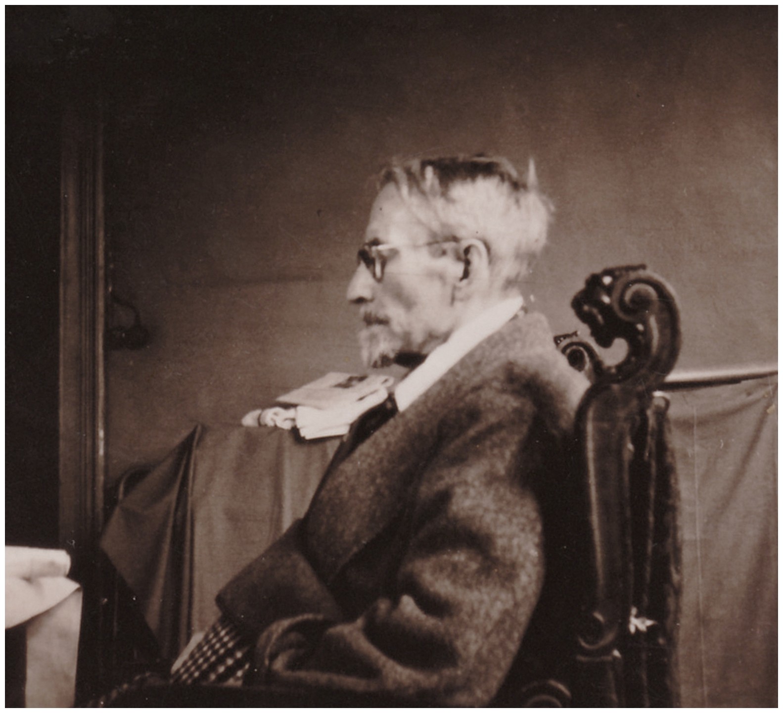 Black-and-white portrait photograph of Karl Kollwitz, shown as an older bearded man wearing glasses in profile. He is seated in a carved wooden armchair, dressed in a dark jacket over a light shirt, and looks to the left. In the background, a table with papers and a dark curtain are visible, suggesting an interior study or office setting.