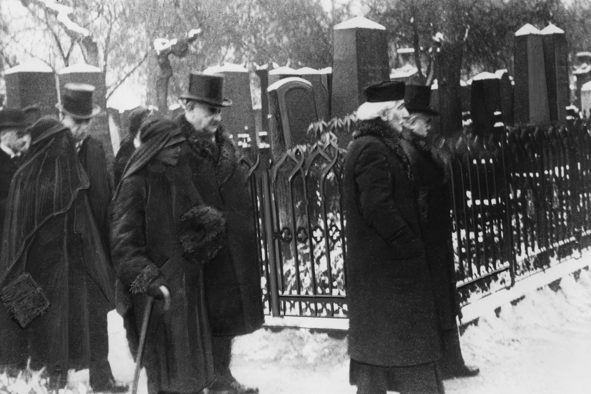 Historic black-and-white photograph showing a group of mourners in dark winter clothing walking along an ornate iron cemetery fence in snowy weather. In the foreground, several figures wear long coats and hats, including a woman with a walking stick and a man wearing a top hat. Large snow-covered gravestones rise in the background, reinforcing the solemn atmosphere of mourning and remembrance.