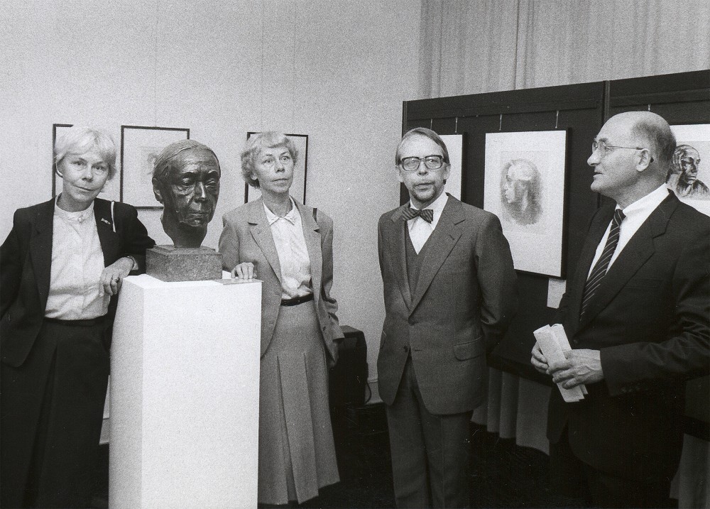 The black-and-white photograph shows Kollwitz’s grandchildren – Jutta Bohnke-Kollwitz, Jördis Erdmann, Arne Andreas Kollwitz and Hans-Joachim Möhle, Chief Executive Officer of the Kreissparkasse Köln (from left) – standing at the opening of the Kollwitz Museum Köln in 1985. Between the granddaughters stands the artist’s bronze self-portrait, with further works visible in the background.