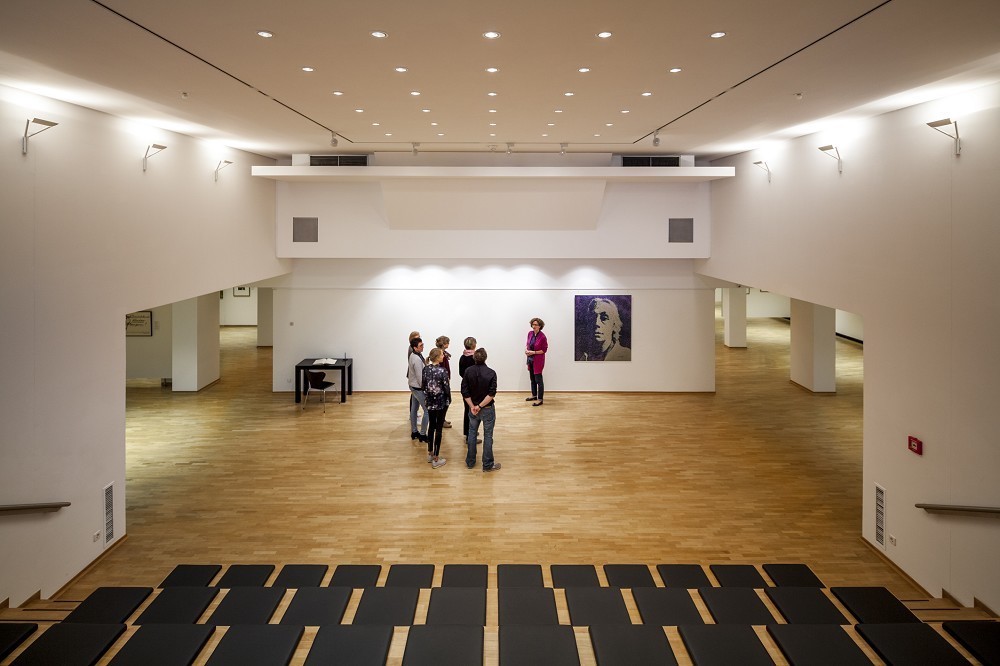 The colour photograph shows a view of the forum at the Kollwitz Museum Köln in 2015: a spacious, bright room with light-coloured wooden flooring; on the display wall hangs a portrait of Kollwitz, reworked by the artist Herbert Döring-Spengler. In the foreground is a group of people. 