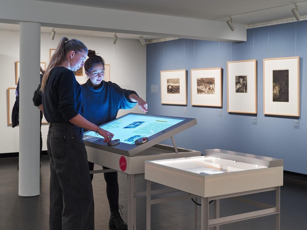The photograph shows a view of the collection presentation for the reopening of the Kollwitz Museum Köln in 2025, featuring the multimedia station on intaglio printing. Two museum visitors are using the desk-like, computer-assisted art education tool.