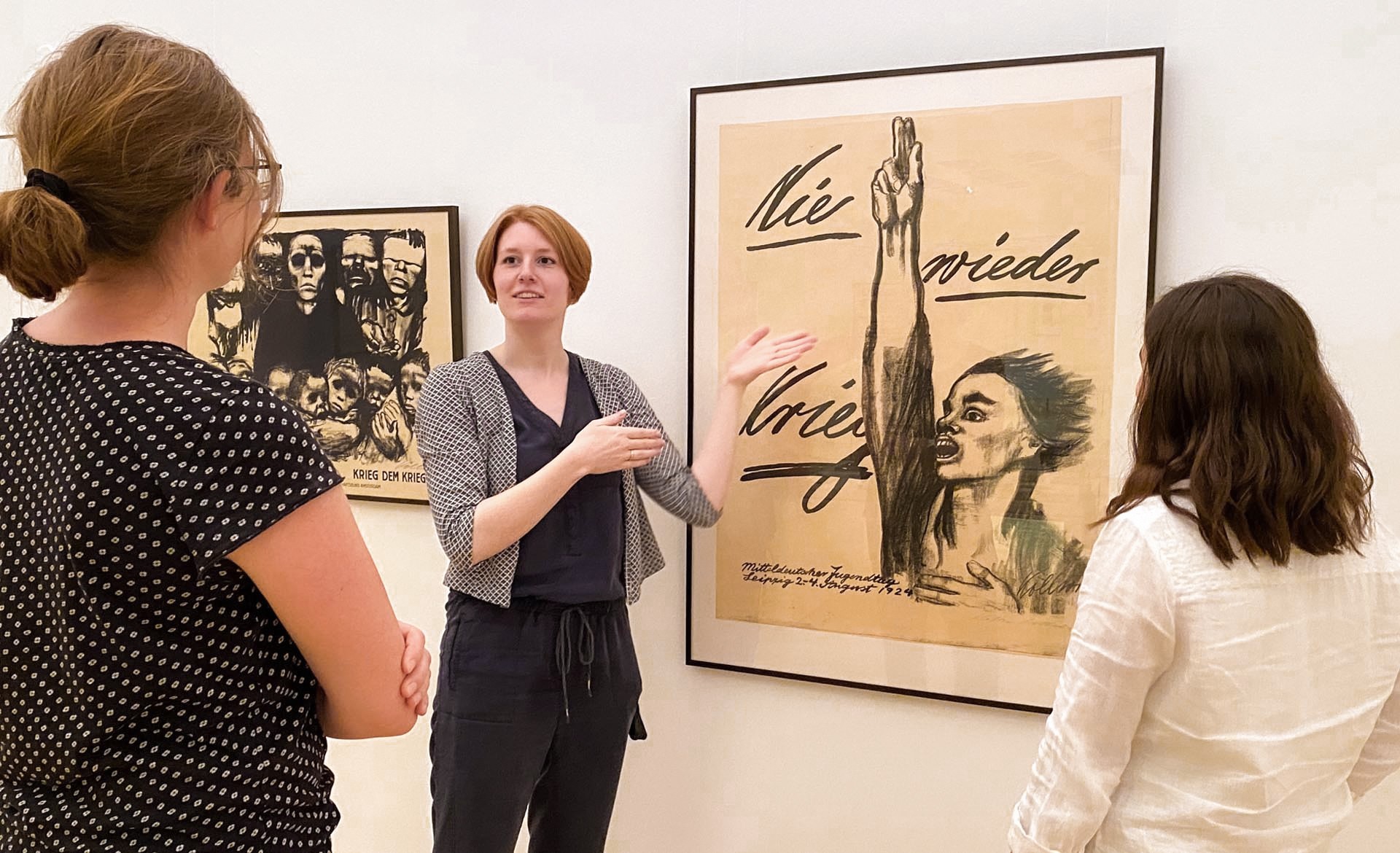 In the brightly lit exhibition rooms of the Kollwitz Museum in Cologne, an art educator uses sweeping hand gestures to explain Käthe Kollwitz’s famous anti-war poster “Never Again War” to two visitors who are listening intently. The artwork takes center stage, featuring an expressive black-and-white charcoal drawing of a person passionately demanding “Never Again War” with her mouth wide open and her hand raised in a vow.