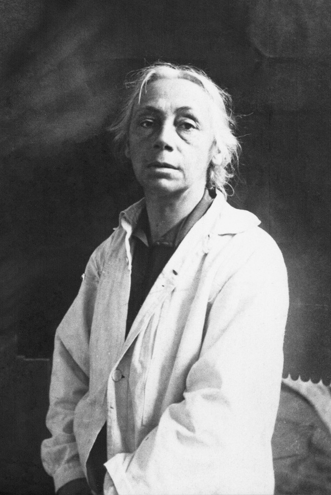 Das Schwarz-Weiß-Porträt zeigt Käthe Kollwitz um 1920, stehend, in einer frontalen Halbkörperaufnahme vor einem dunklen, unbestimmten Hintergrund. Die etwa 53-jährige Künstlerin trägt einen hellen, schlichten Arbeitskittel, der über der Brust leicht geöffnet ist und den Blick auf ein dunkleres Untergewand freigibt. Ihr graues Haar ist am Oberkopf gescheitelt und an den Seiten locker zurückgenommen, wobei einzelne Strähnen ihr Gesicht umrahmen. Ihr Blick ist direkt in die Kamera gerichtet und wirkt ernst. Fotograf:in unbekannt. Ein Reproabzug dieses Fotos aus dem Nachlass der Künstlerin befindet sich im Archiv des Kollwitz Museum Köln, Inv.: Nr. F 01.b-12.