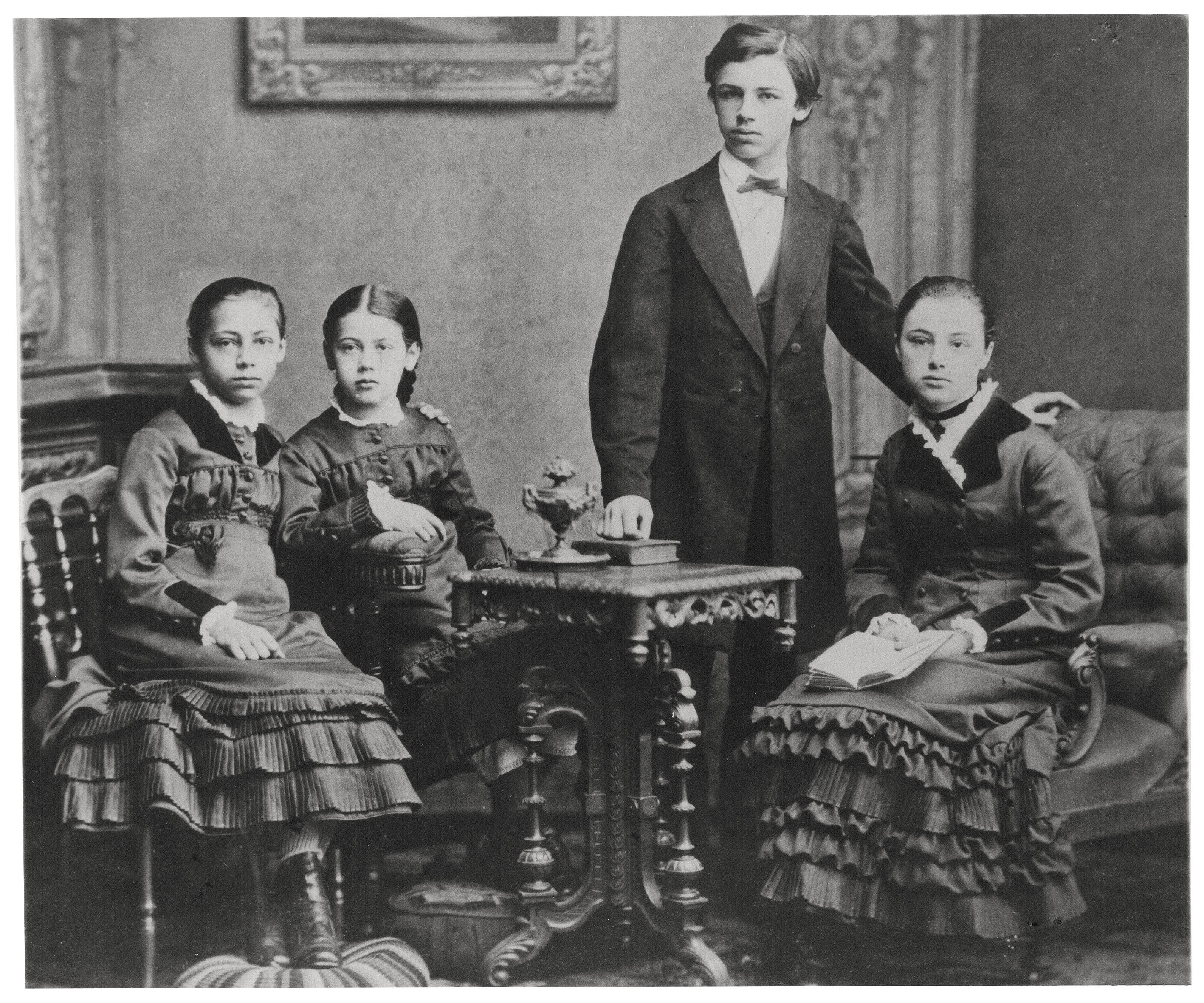 Das Bild ist eine Schwarz-Weiß-Fotografie aus den Jahren um 1880, das die vier vornehm gekleideten Geschwister Schmidt, von links: Käthe, Lisbeth, Konrad, Julie, in einem bürgerlichen Wohnzimmer zeigt. Im Zentrum steht Konrad in dunmklem Anzug mit weißem Hemd und Fliege, umgeben von den drei Mädchen in dunklen Kleidern mit auffälligen Rüschen und weißen Kragen. Die Gruppe posiert ernst und unbeweglich um einen kunstvoll verzierten Holztisch.