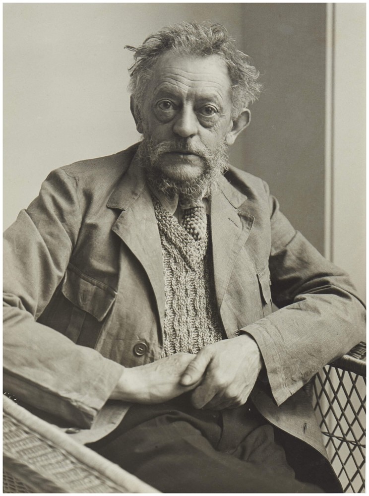 Das Schwarz-Weiß-Foto zeigt den Bildhauer Ernst Barlach als Brustporträt in einer frontalen Ansicht, fotografiert 1933 von Hildegard Heise. Er ist ein älterer Mann mit zerzaustem, hellem Haar, einem dichten, graumelierten Vollbart und einem intensiven, direkt in die Kamera gerichteten Blick. Barlach trägt eine grobe, helle Jacke über einer markant gestrickten Weste mit Krawatte und hält seine Hände, die Ellbogen auf den Lehnen seines Korbsessels, über seinem Schoß.
