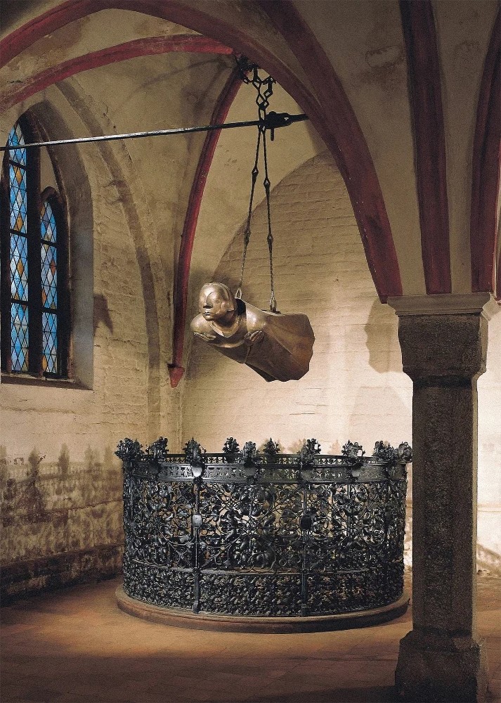 Das Foto das Fotografen Uwe Seemann zeigt die Bronzeplastik »Der Schwebende« von Ernst Barlach, den Drittguss von 1952 nach dem Kölner Exemplar, an seinem heutigen Ort, dem ursprünglich vorgesehenen Platz im östlichen Joch des Nordschiffes im Dom zu Güstrow. Die lebensgroße, waagerecht schwebende Bronzefigur hängt an zwei Ketten frei in der Luft, wobei ihr gerader Körper und die vor der Brust gekreuzten Arme eine tiefe Ruhe ausstrahlen. Das Gesicht der Skulptur hat geschlossene Augen und wirkt wie insich versunken, während die fließende Form des Gewandes den Eindruck eines sanften Fluges verstärkt. Unter dem „Schwebenden“ steht ein kreisrundes, kunstvoll verziertes Schmiedeeisengitter, das die Figur zusätzlich im Raum verankert und einen Ort des Gedenkens markiert.
