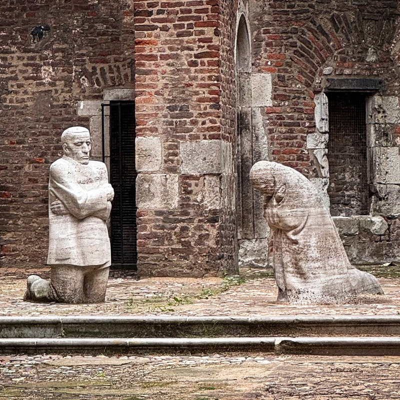 Das Foto von Christian Nitz zeigt das Mahnmal »Trauernde Eltern« von Käthe Kollwitz, das im offenen Innenraum der Kirchenruine Alt St. Alban in Köln aufgestellt ist. Inmitten der verwitterten Backsteinmauern knien sich zwei lebensgroße, blockhaft massive Steingestalten in stillem Schmerz gegenüber: Links verharrt der Vater mit starr geradeaus gerichtetem Blick und eng vor der Brust verschränkten Armen, während rechts die Mutter tief gebeugt und in sich gekehrt um den gemeinsamen Sohn trauert.