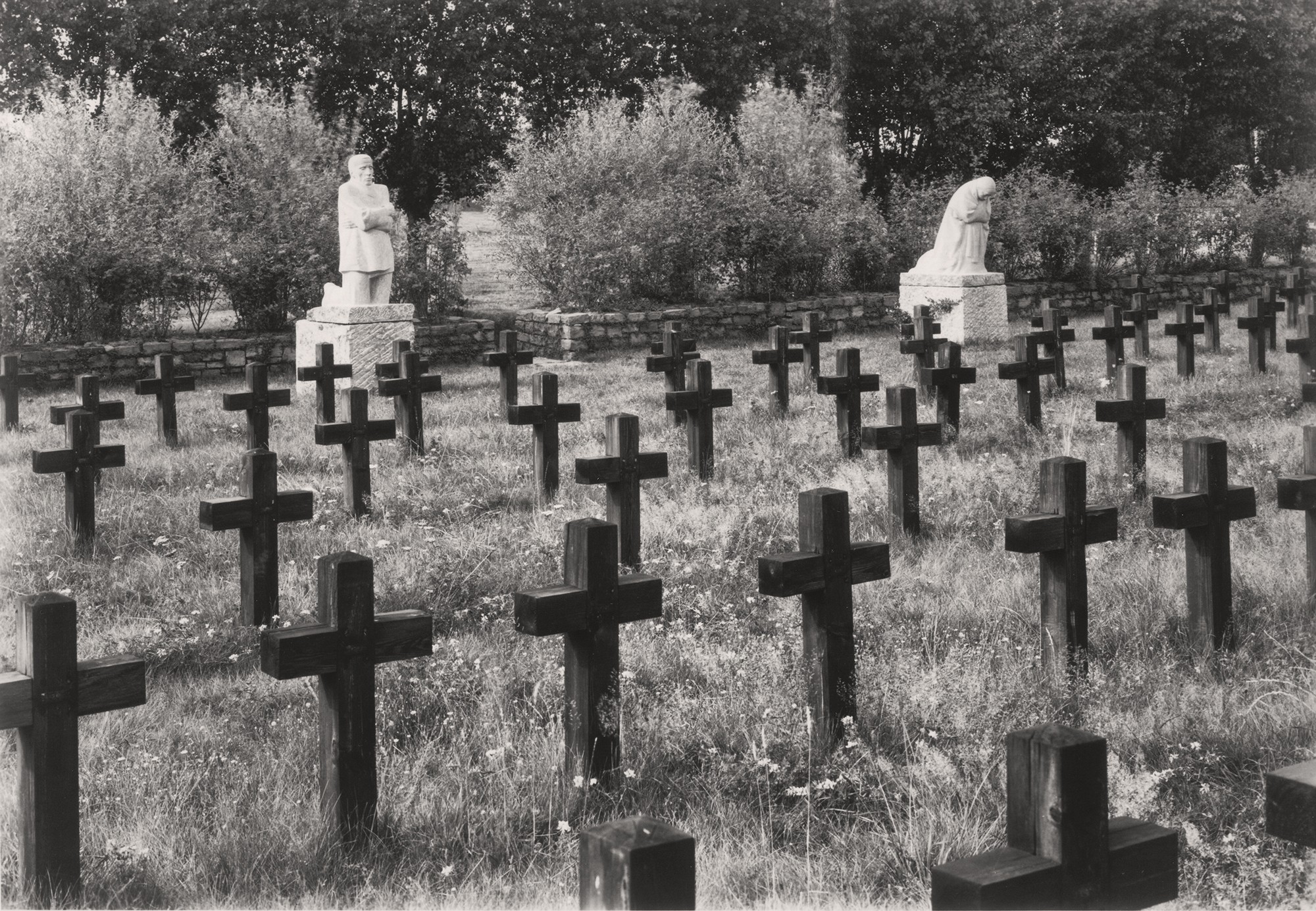 Die Schwarz-Weiß-Fotografie von Willi Moegle aus dem Jahr 1937 zeigt das Skulpturenpaar »Trauernde Eltern« in seiner originalen Aufstellung von Juli 1932 bis 1955 auf dem Soldatenfriedhof Roggeveld in Belgien. Mit einem Blick von rechts ist im Vordergrund eine sommerliche Wiese mit zahlreichen Holzkreuzen zu sehen, im Hintergrund vor einer niedrigen Bruchsteinmauer die Skulpturen - rechts der Vater, links die Mutter - zwischen beiden öffnet sich die Steinmauer als schmaler Ausgang aus dem Friedhof.