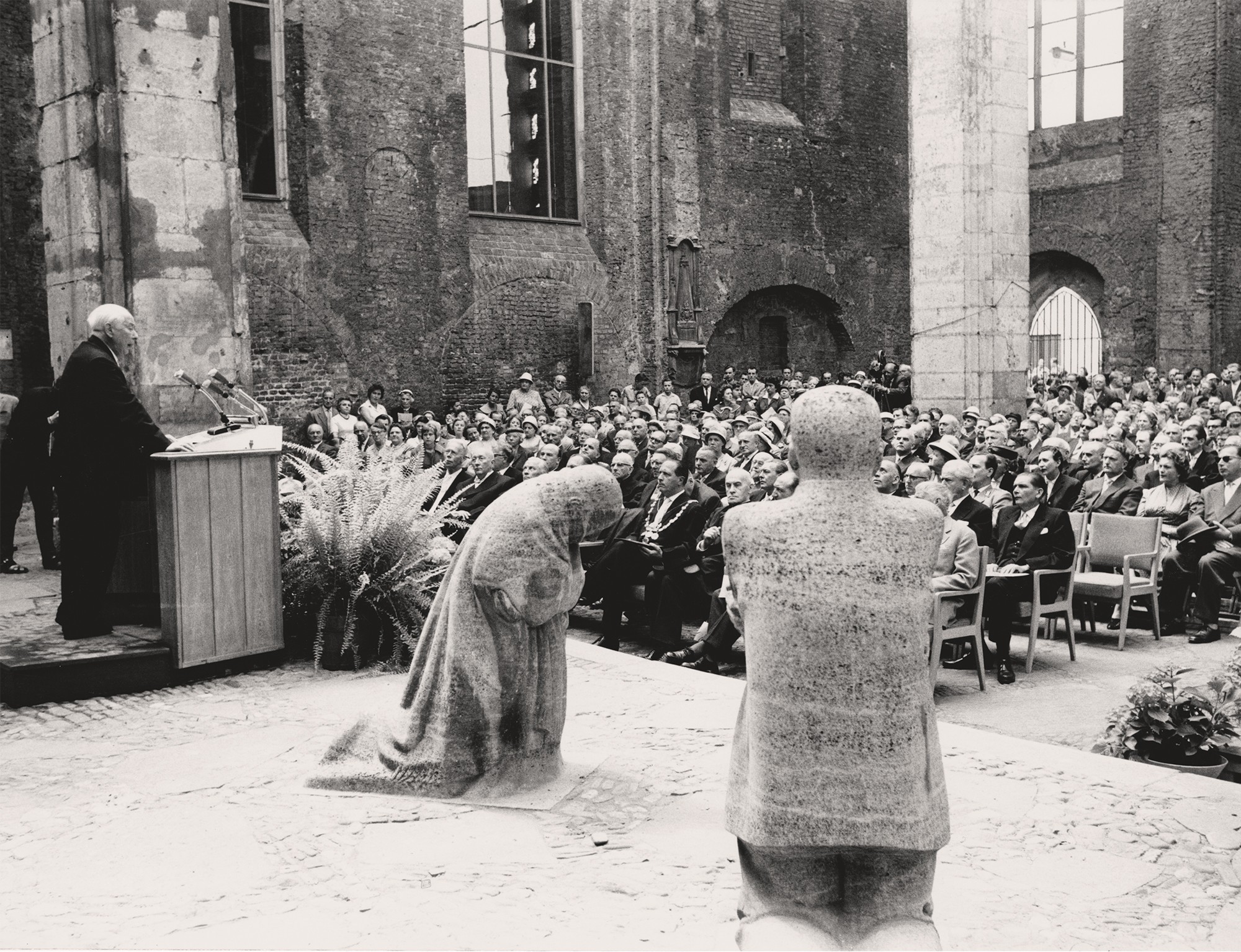 Die Schwarz-Weiß-Fotografie von Hansherbert Wirtz zeigt einen Blick in die Kölner Kirchenruine von Alt St. Alban vom Chor aus in das Publikum bei der Einweihung als Bundesgedenkstätte. Zu sehen sind die beiden Steinskulpturen »Trauernde Eltern«, der Vater links, die Mutter mittig, rechts am Bildrand Bundespräsident Theodor Heuss während seiner Rede.