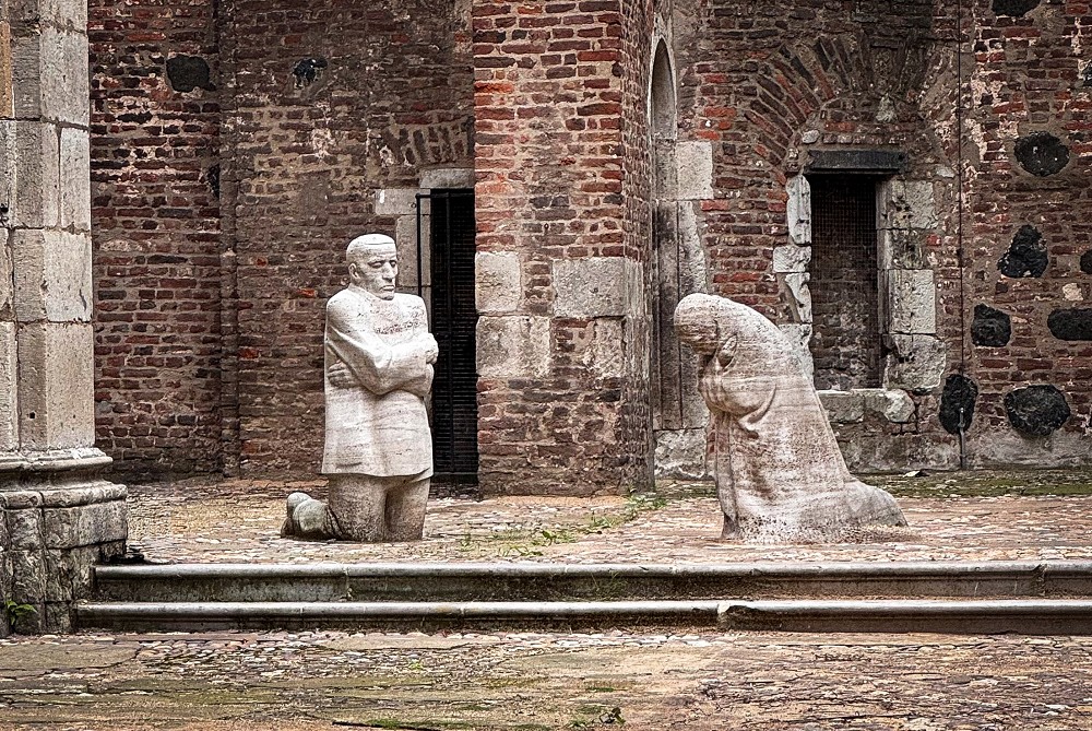Das Foto von Christian Nitz zeigt das Mahnmal »Trauernde Eltern« von Käthe Kollwitz, das im offenen Innenraum der Kirchenruine Alt St. Alban in Köln aufgestellt ist. Inmitten der verwitterten Backsteinmauern knien sich zwei lebensgroße, blockhaft massive Steingestalten in stillem Schmerz gegenüber: Links verharrt der Vater mit starr geradeaus gerichtetem Blick und eng vor der Brust verschränkten Armen, während rechts die Mutter tief gebeugt und in sich gekehrt um den gemeinsamen Sohn trauert.