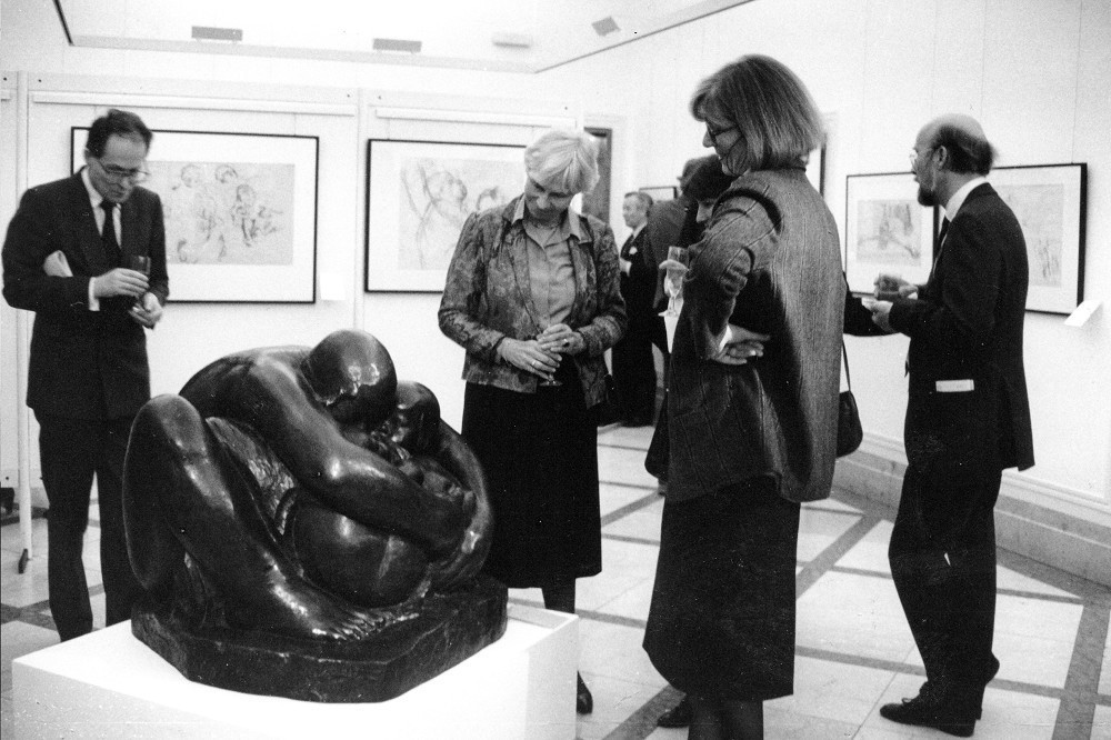 Das Schwarz-Weiß-Foto zeigt einen Blick in die ersten Ausstellungsräume des Museums auf der ehemaligen Vorstandsetage der Kreissparkasse Köln, 1986:  Mehrere Frauen und Männer betrachten Werke der Kollwitz, im Zentrum die große Bronzeplastik »Mutter mit zwei Kindern«. 
