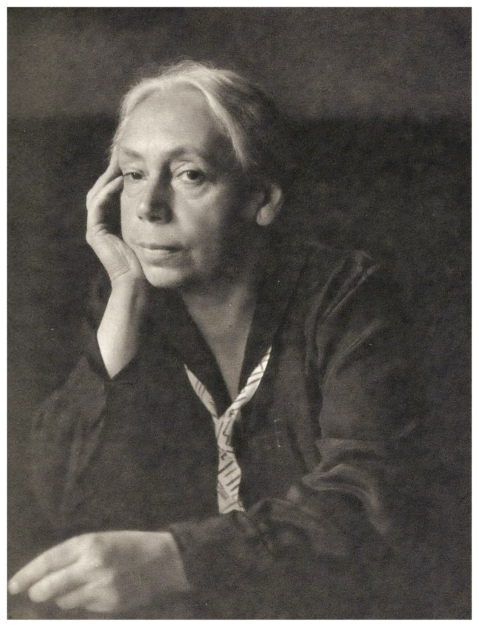 Das Schwarz-Weiß-Porträt zeigt Käthe Kollwitz mit hellem, nach hinten gekämmtem Haar in einer nachdenklichen Pose. Sie blickt mit ernstem, melancholischem Ausdruck leicht am Betrachter vorbei, während sie ihren Kopf auf ihre rechte Hand stützt. Bekleidet ist sie mit einer dunklen Bluse und einer hellen, gemusterten Krawattenschleife vor einem ruhigen, dunklen Hintergrund.