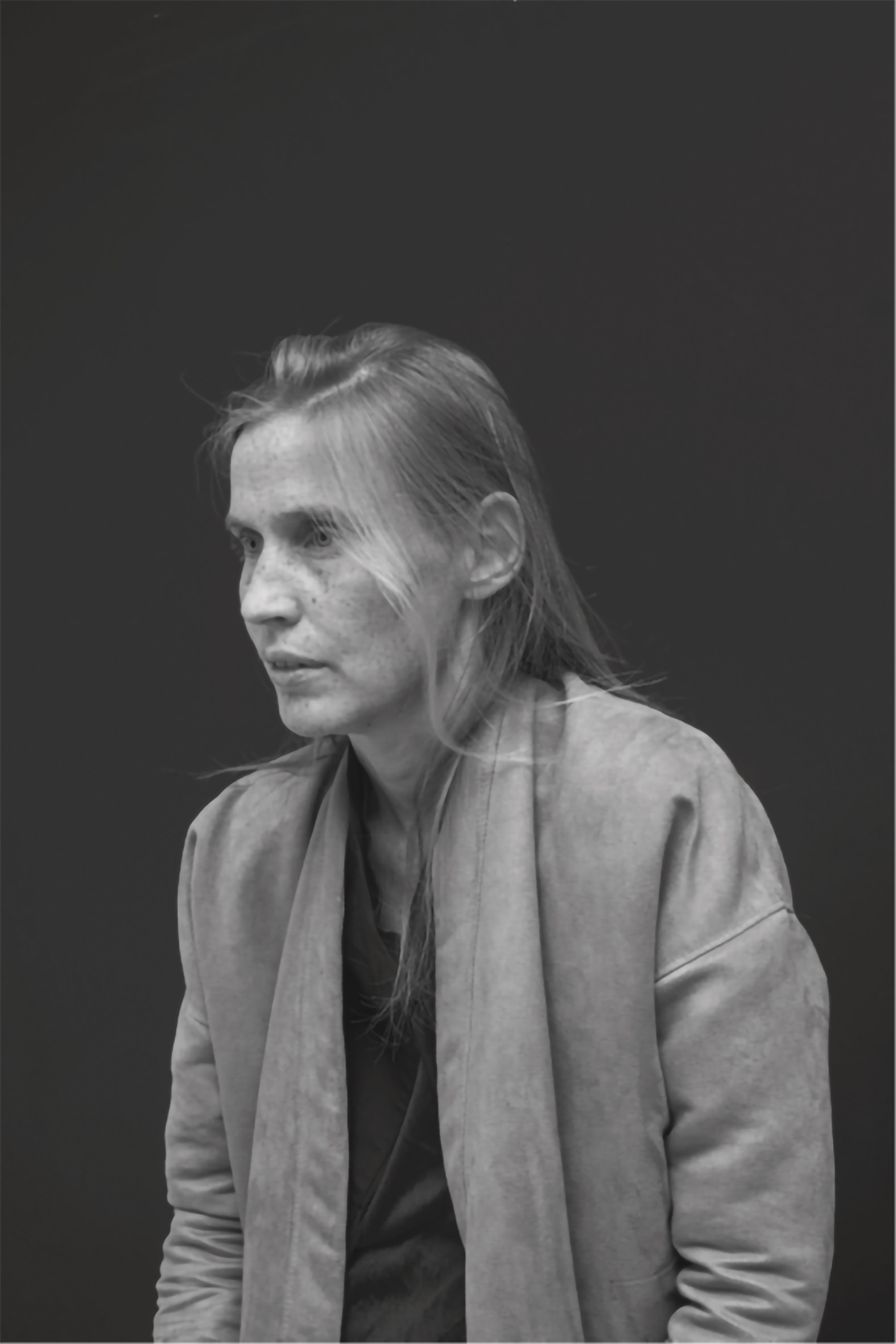 This black-and-white portrait shows a woman with long, light-coloured hair, viewed in three-quarter profile against a neutral, dark background, looking to the left. She has a pensive or serious expression and is wearing an open, softly draping jacket over a dark top. The light falls on her face from the front, gently highlighting her delicate features and the texture of her clothing.