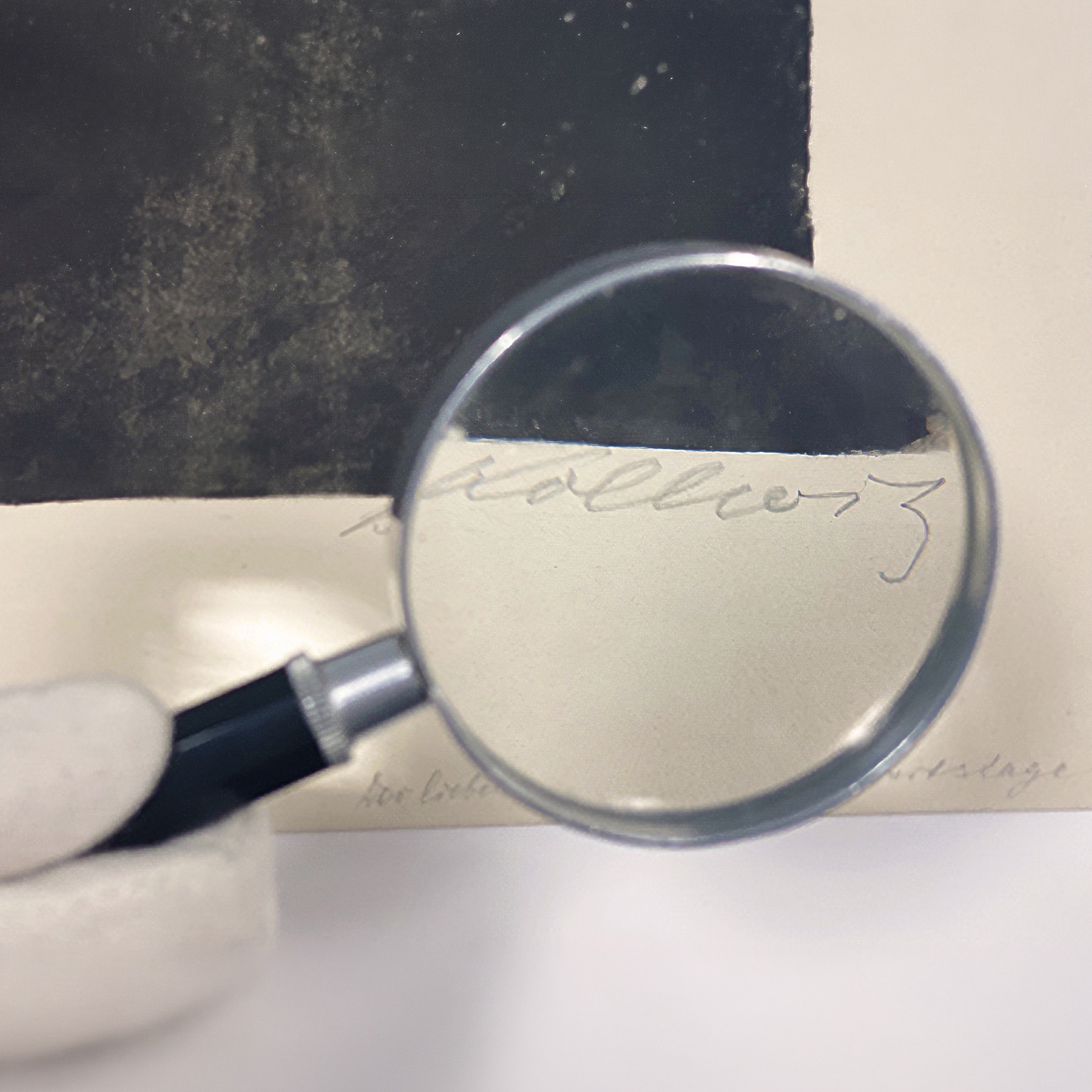 Das Bild zeigt eine Lupe, die von einer Hand in einem weißen Handschuh über die handschriftliche Signatur „Kollwitz“ auf einem hellen Papierrand gehalten wird. Durch die Vergrößerung ist der Namenszug der Künstlerin unterhalb eines dunklen, texturierten Kunstwerks deutlich lesbar.