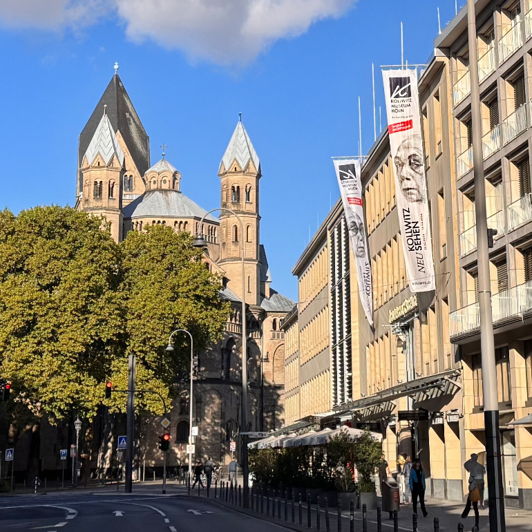 Das Bild zeigt die moderne, helle Fassade des Käthe Kollwitz Museums am belebten Kölner Neumarkt, von der lange vertikale Fahnen mit dem Motiv aktuellen Sonderausstellung herabhängen. Im Hintergrund ragen die markanten, grauen Türme der romanischen Basilika St. Aposteln hinter grünen Baumkronen in den blauen Himmel.